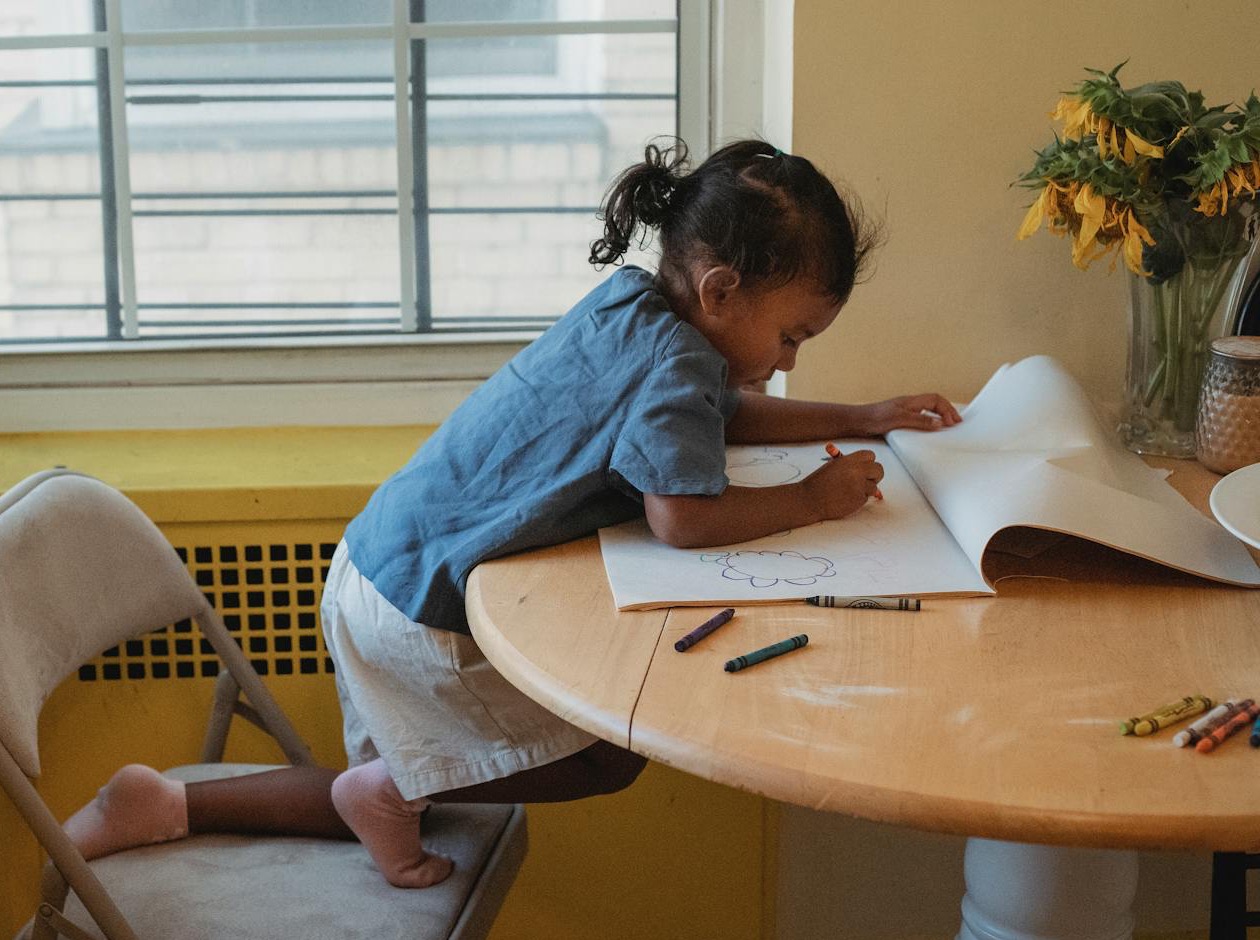 A child drawing at a table by a window.